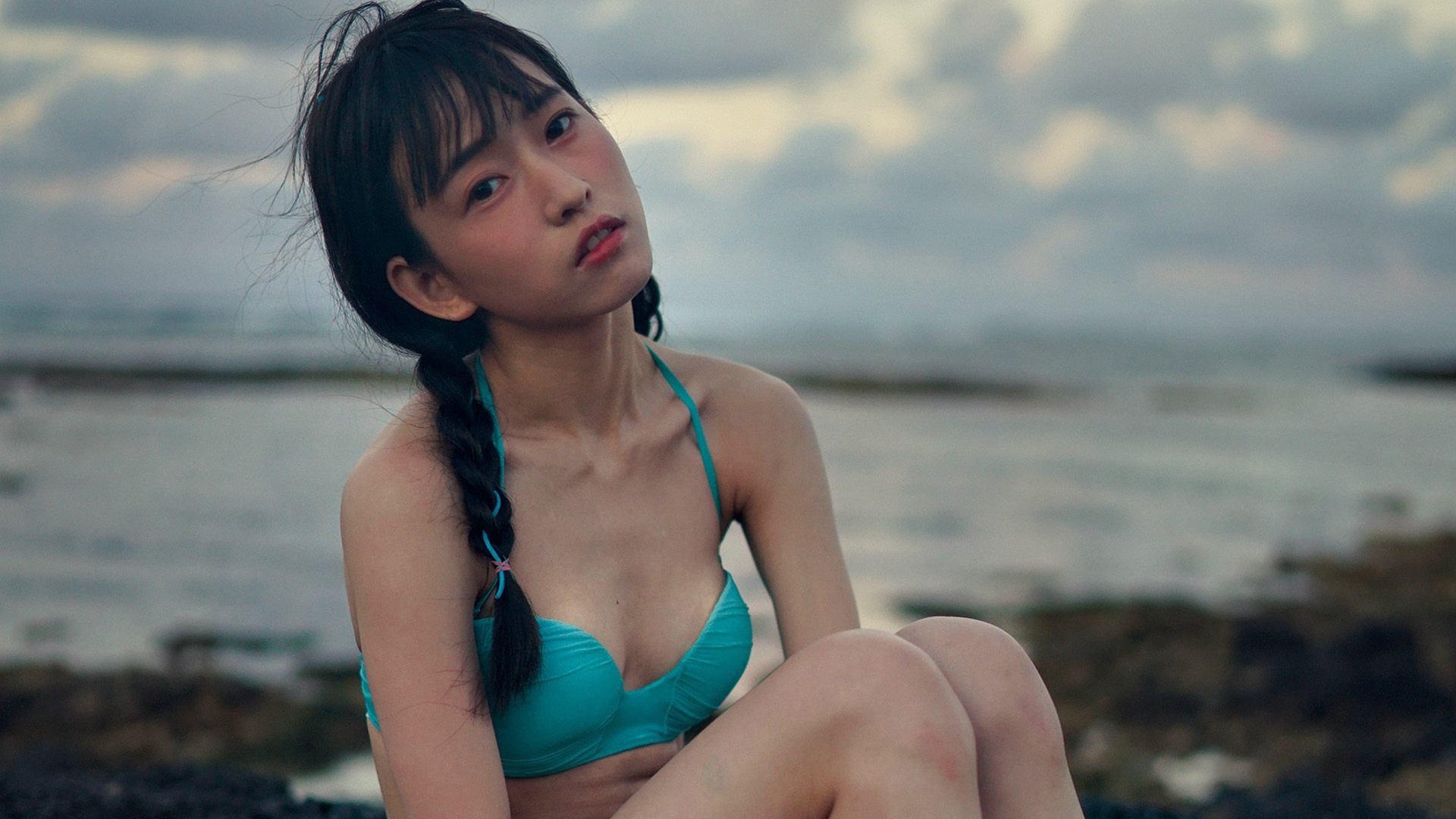 穿青绿色比基尼的麻花辫少女坐在海边礁石上望向镜头