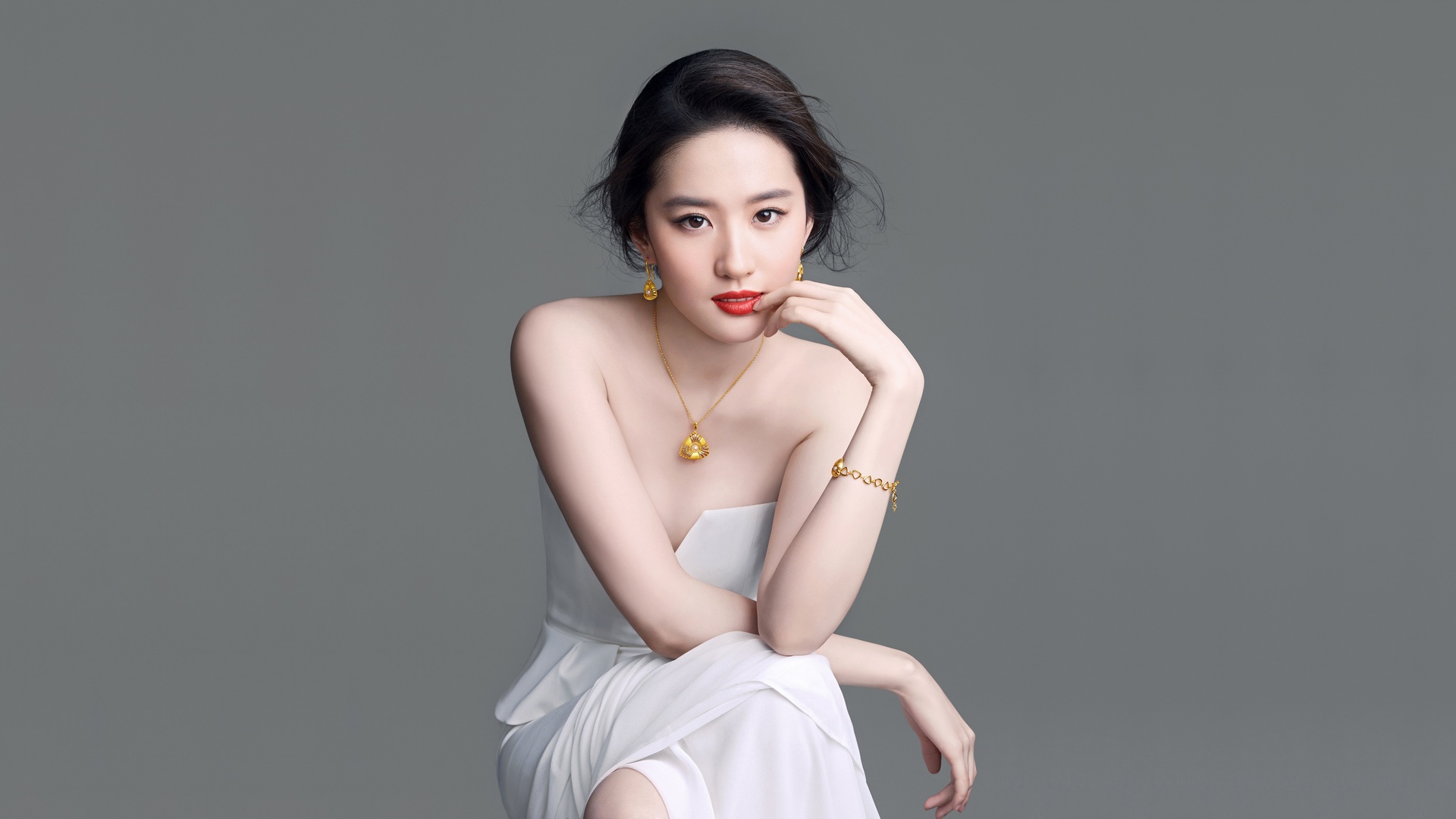 影棚内一名黑色盘发的东亚女性，涂红色口红，佩戴全套金色珠宝首饰，身穿白色抹胸开叉长裙，坐姿朝向镜头，背景为纯灰色无装饰墙面。