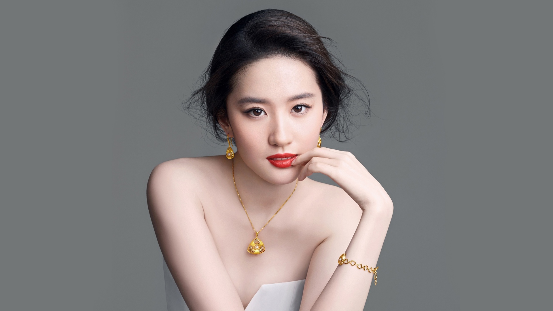 浅灰色纯色背景前，盘发年轻东亚女性身着白色抹胸，佩戴嵌珍珠黄金耳环、项链与手链，涂正红色唇妆，面向镜头正视。