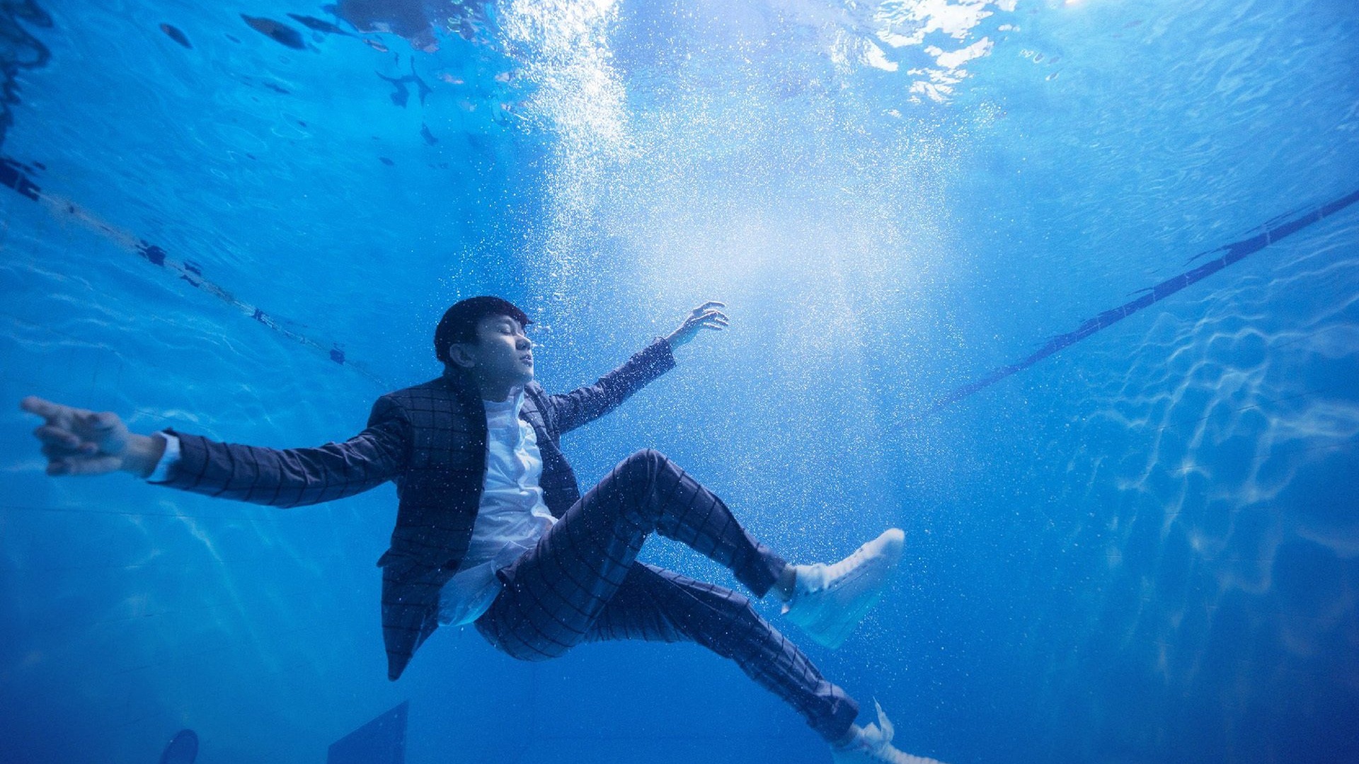 身着深色格纹西装的短发男子闭眼漂浮在蓝色水下，双臂向两侧伸展，水中遍布气泡，光线从水面射入形成光束，背景可见泳池泳道线。