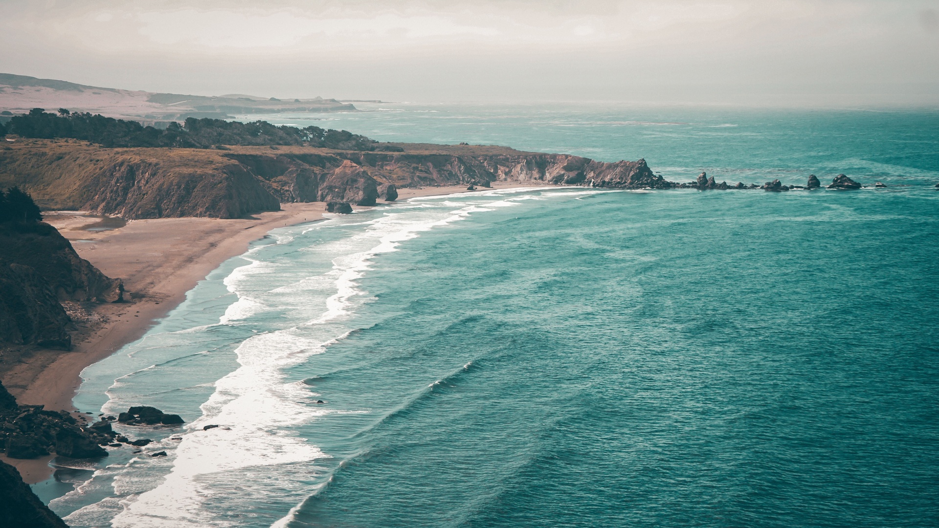 俯瞰视角的滨海海湾风景，青蓝色海面拍打着弧形浅棕沙滩，沿岸分布海蚀崖与近海礁石，整体为低饱和复古调色。