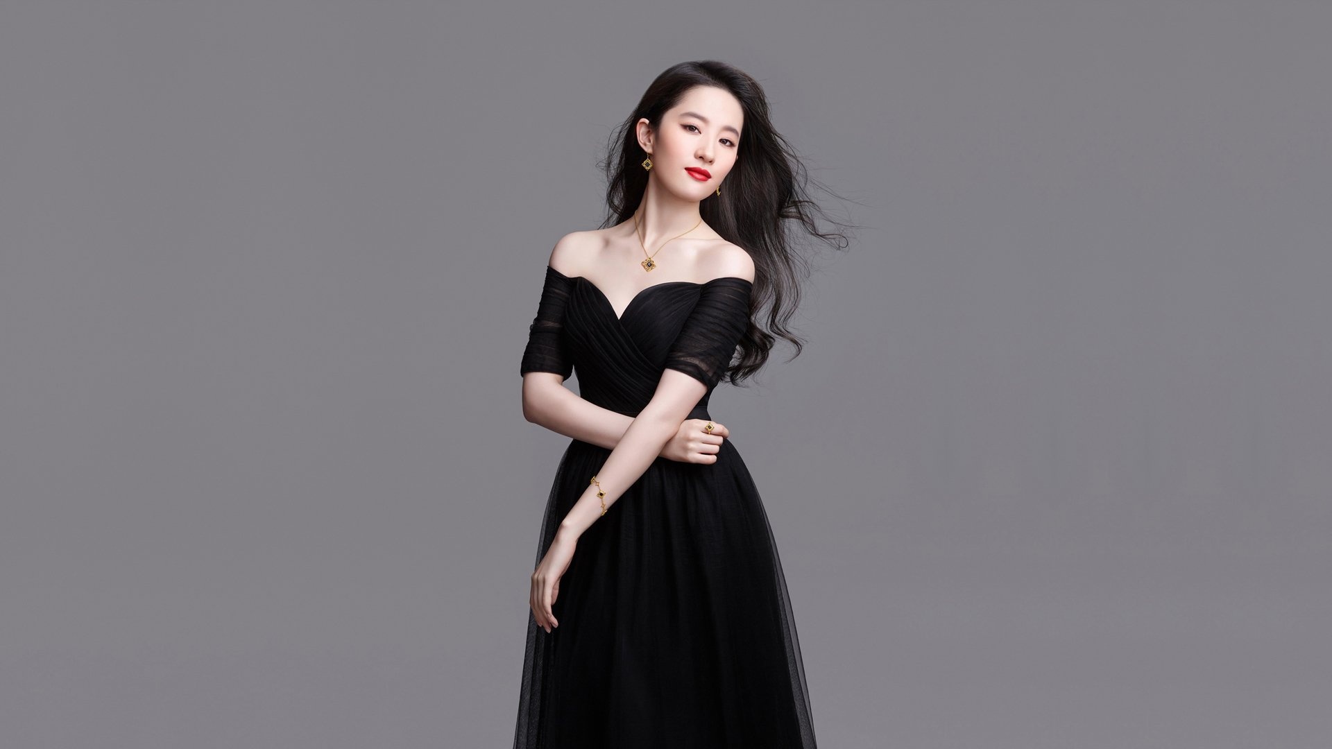 灰调纯色影棚背景前，一名留黑色长卷发、涂红色口红的女性，身着黑色露肩纱质长裙，佩戴带深色宝石的金色首饰，姿态优雅地正对镜头站立。