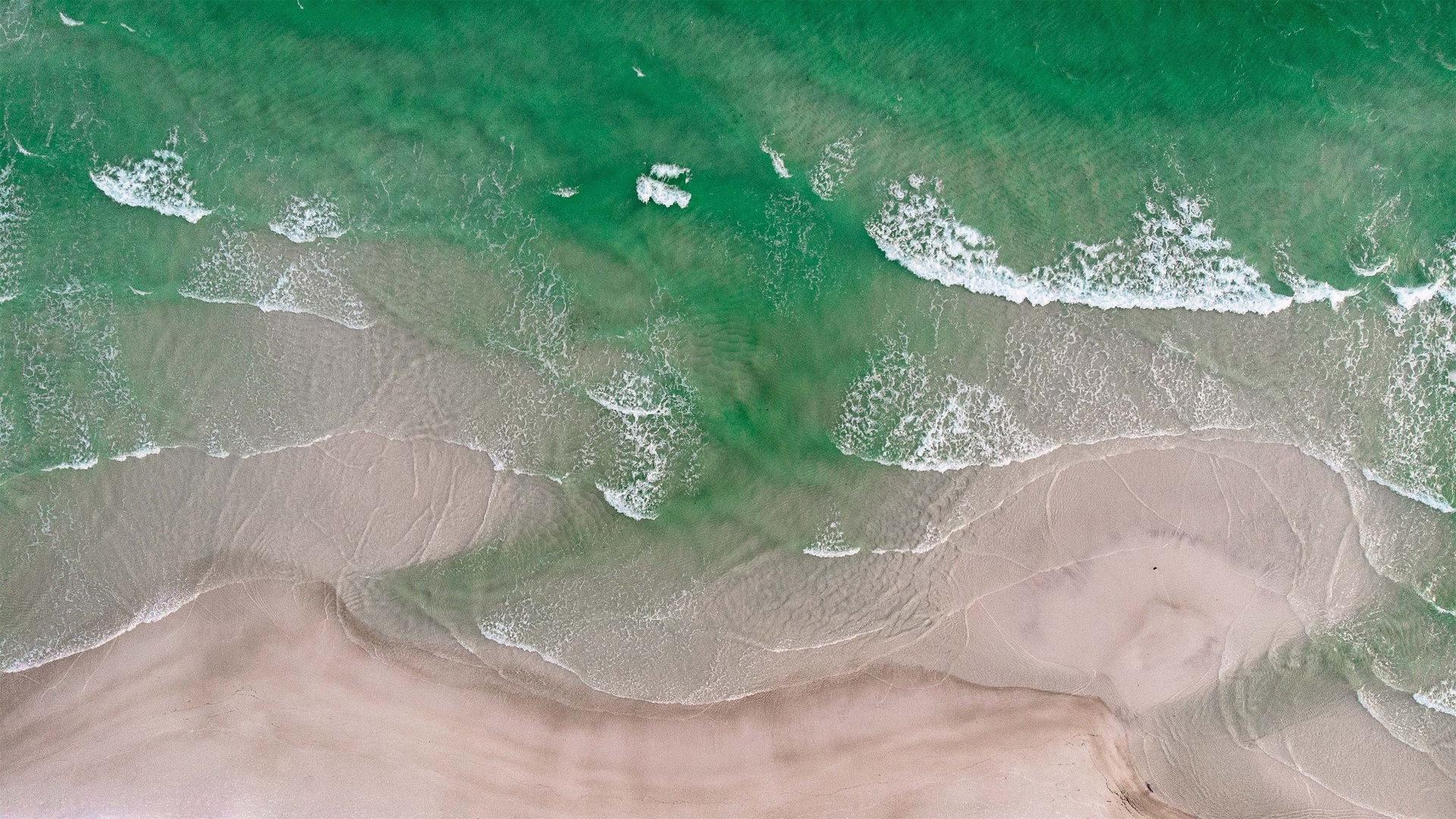 正俯视航拍视角下，青绿色澄澈海浪涌向浅米色沙滩，海面遍布白色碎浪泡沫的海滨实景。