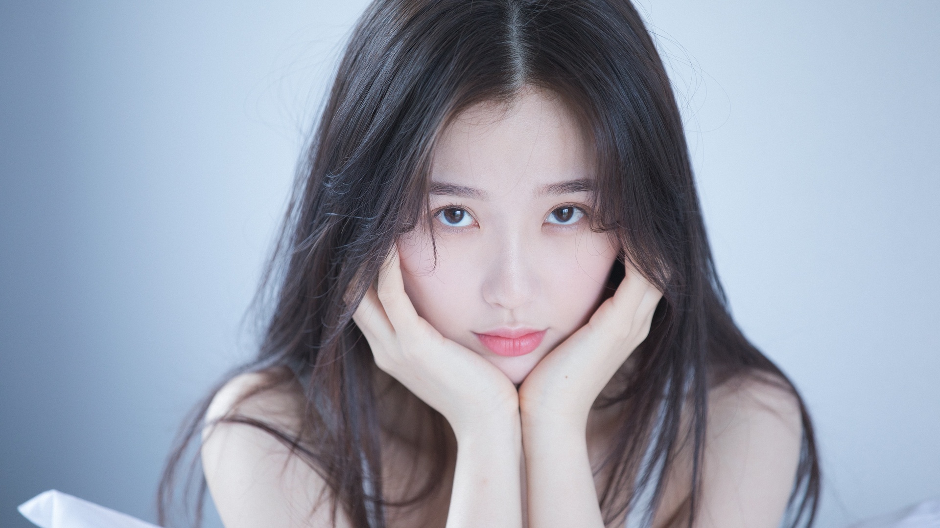浅灰纯色背景的室内人像，一位深色长发女性双手托住脸颊，面向镜头，清透淡冷色调，风格简约柔和。