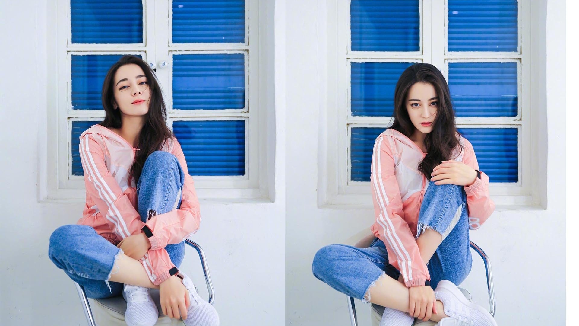 黑色长卷发年轻女性身着粉色白条纹连帽运动外套与毛边牛仔裤，坐于金属座椅上，背景为带蓝色百叶窗的白色分格木窗与白色墙面。
