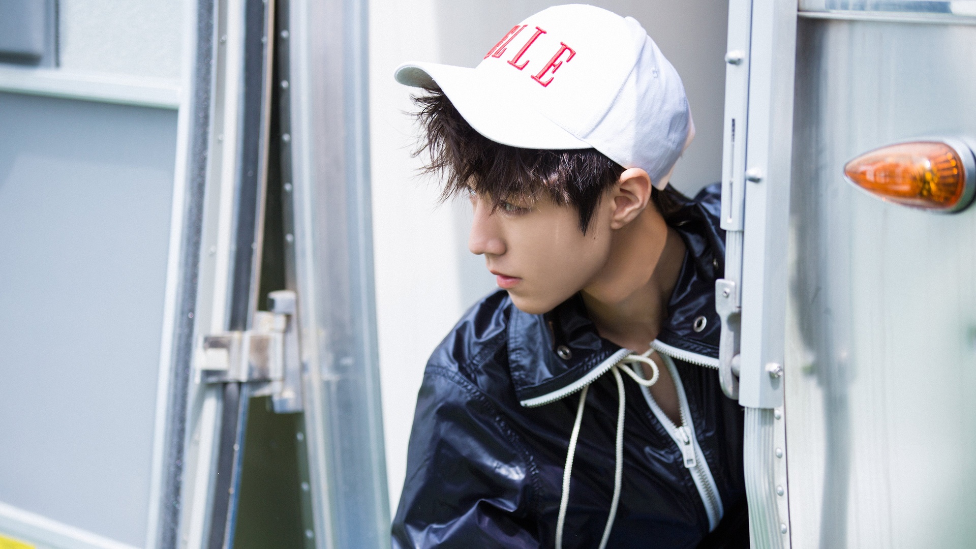 tfboys主题画面中，戴白棒球帽、穿藏蓝亮面连帽外套的年轻男性侧立于银色金属房车车门旁，背景可见车门金属锁扣与橙色转向灯。