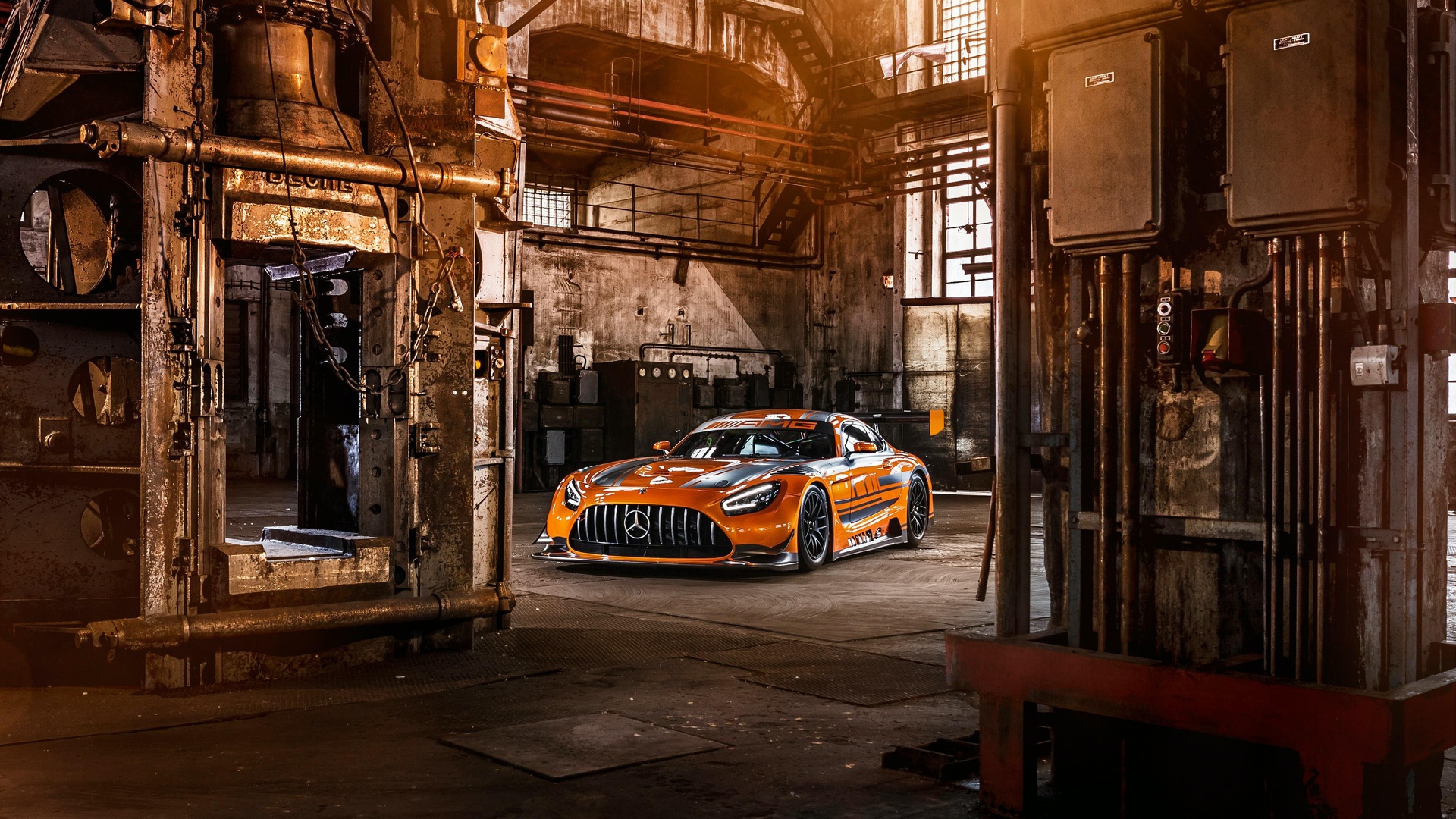 废弃工业厂房内停放着橙黑涂装的奔驰AMG GT3赛车，车头带有三叉星标识，周围可见锈蚀金属机械、管道与配电箱，暖金色光线笼罩场景。