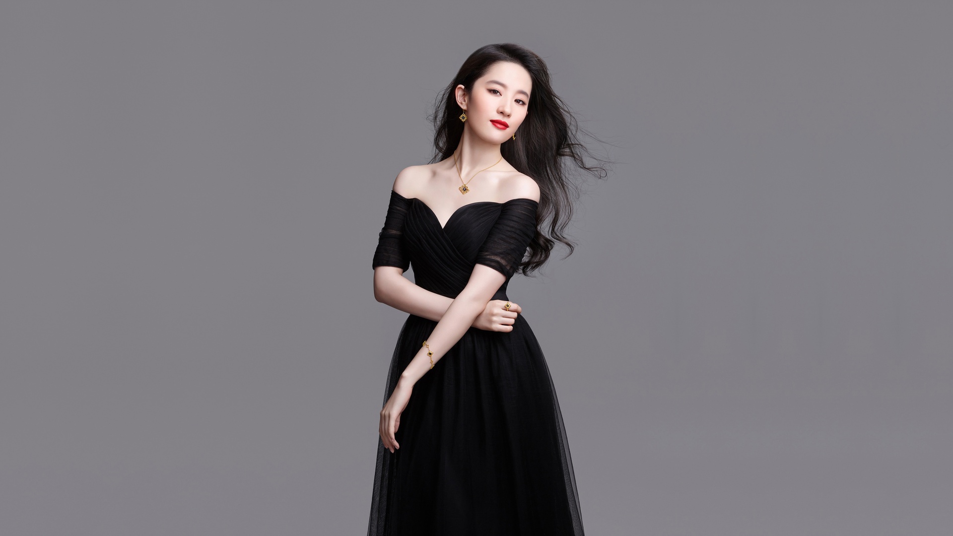浅灰影棚中，深色长卷发女性身着黑色露肩网纱连衣裙，佩戴成套金色嵌宝石珠宝。