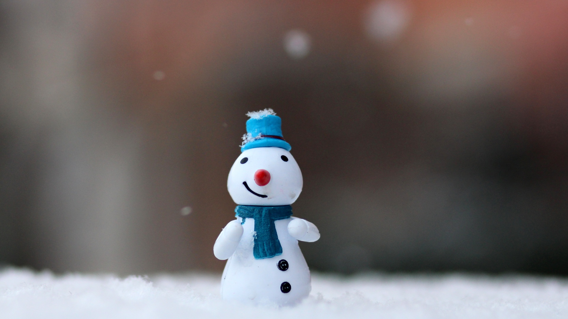 冬日雪面近景中，戴蓝色礼帽、同色围巾的小雪人摆件立于白积雪地上，背景为棕褐色虚化散景。