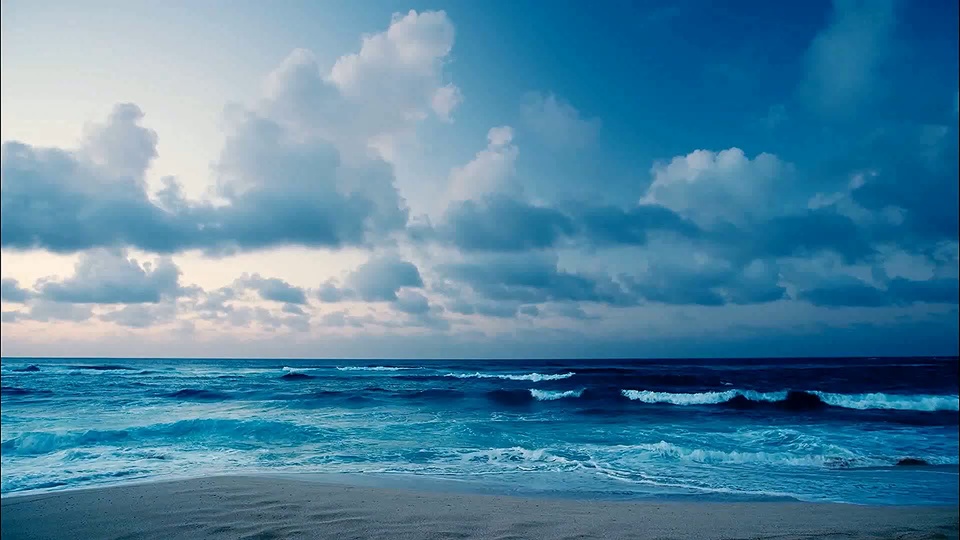 从近岸视角拍摄的滨海海景，近侧是浅米色沙滩，前方是泛着白色浪边的青蓝色海面，上方是从靠近海平线的浅灰蓝向上过渡到深邃深蓝的天空，空中散落着蓬松的白色云团。