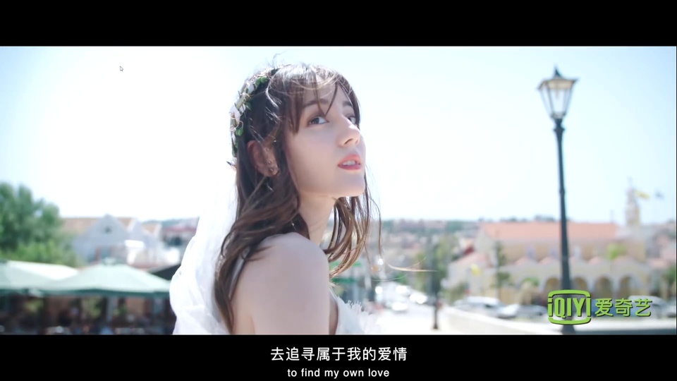 晴朗浅蓝色天空下，身着白色婚纱、头戴绿叶花卉头饰的年轻女性侧首望向画面右上方，背景是虚化的欧式临街建筑群、黑色复古路灯与行道树。