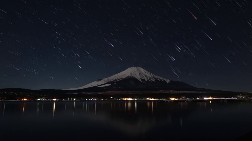夜色星空下，覆雪的锥形山体矗立在平静湖畔，深黑夜空铺展斜向星轨，山体与岸边暖黄灯光在湖面形成清晰倒影。