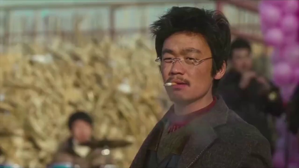 男明星留凌乱黑色短发戴细框眼镜叼香烟，穿深色粗花呢外套配红色领边，站在布满枯黄秸秆的户外集市中，背景可见模糊摊位与紫色气球串。