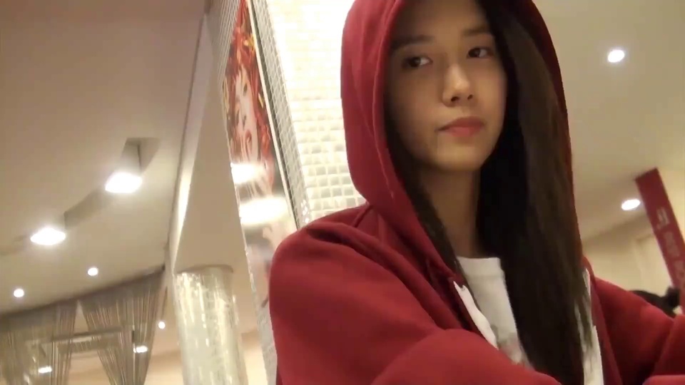 长发年轻女性林允儿身着酒红色连帽卫衣侧身看向画面左侧，背景是带圆形顶灯的室内商业空间，旁有红色边框装饰海报，写实自拍风格，色调偏暖。
