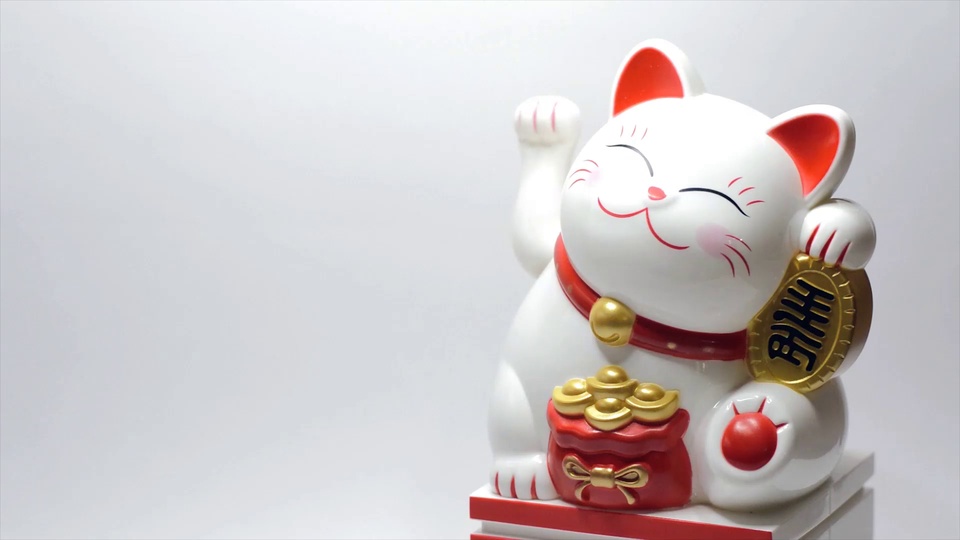 浅灰白色纯色背景上，单爪抬起的白底红内衬招财猫佩戴红项圈金色铃铛，怀抱刻有日文的金牌，旁侧摆放红袋与金元宝，左侧留有大面积空白。