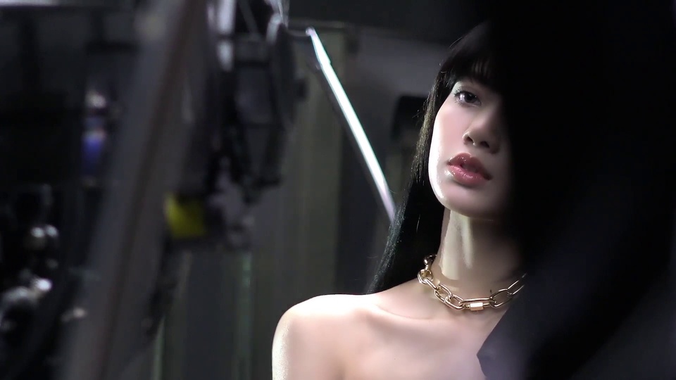 暗调室内摄影布景，留黑色齐刘海长直发、佩戴金色粗颈链的blackpink Lisa裸露肩颈，旁侧斜置长条白色补光灯条，前景有虚化的摄影器械部件