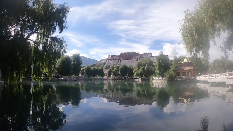实拍湖畔风光，垂柳夹岸，湖面清晰倒映山坡上的宫堡建筑，天空飘着白云