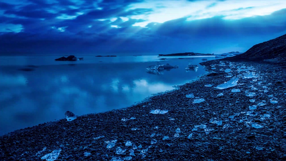 深蓝色调冰岸湖景，黑砾石海滩散落透明碎冰，平静湖面倒映天空与碎冰，远处有暗色低矮岸洲