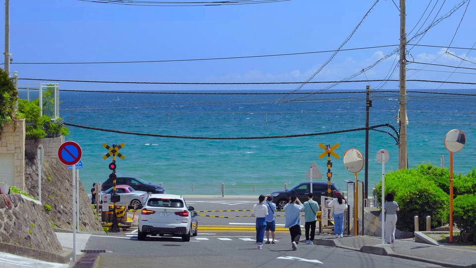 晴朗白日的临海铁道下坡公路，远处是蓝绿渐变的澄澈海面，行人与车辆停靠在道口旁。