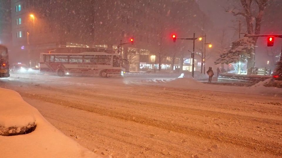 雪夜积雪覆盖的城市马路，路口亮着暖橙色交通信号灯，一辆红白配色的城市公交车行驶在路上，冷白色积雪与暖调灯光形成鲜明色彩对比。