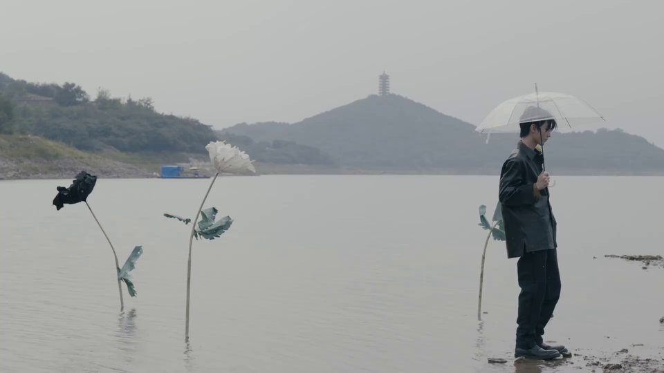 灰调雾感开阔湖景，远处山顶立有古塔，身着深灰外套黑裤的男性持透明伞立于湖边浅滩，湖水中立有三枝大型纸艺花，分白黑两色。