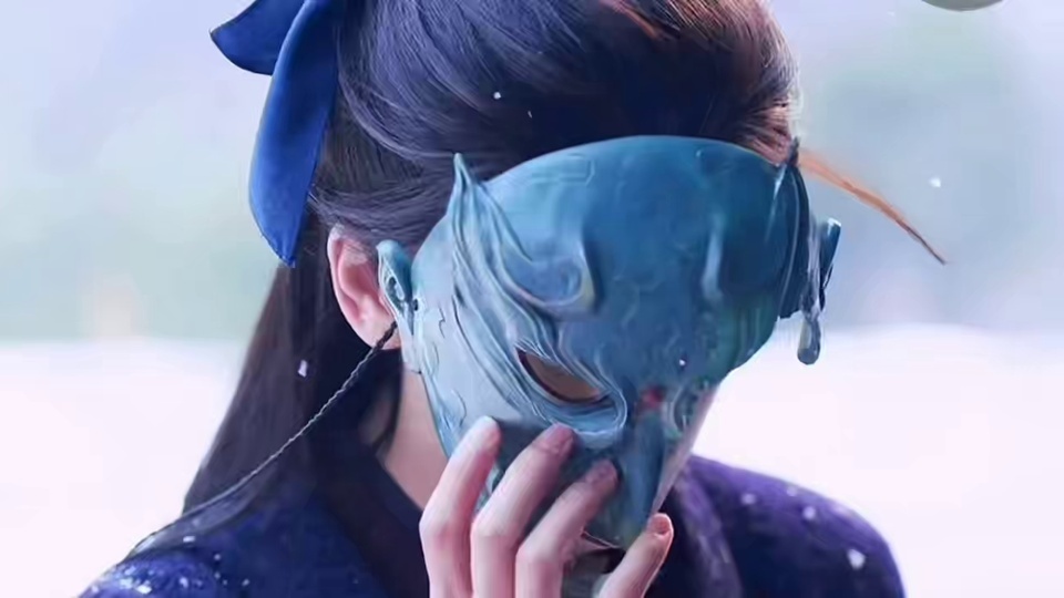 张凌赫身着深紫色带白点古装，束发配深蓝色发饰，手捧青蓝色纹理面具，背景为虚化淡色朦胧外景。