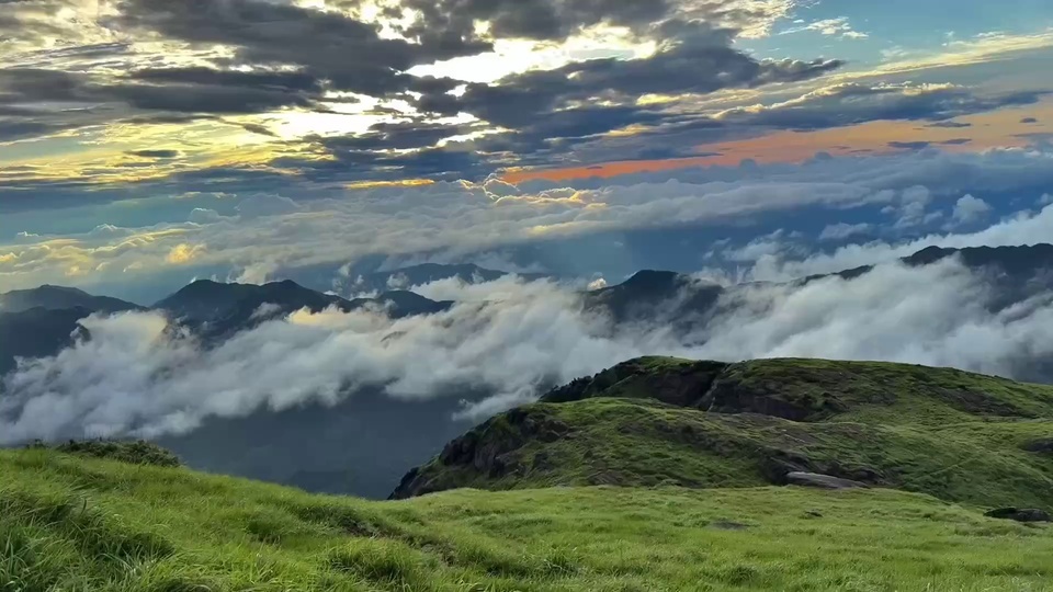 写实山巅自然风景，前景是覆盖鲜绿草被的起伏山地，中后区域铺展亮白色层叠云海，远处露出远山轮廓，天空呈现暖橙向灰蓝过渡的柔和渐变。
