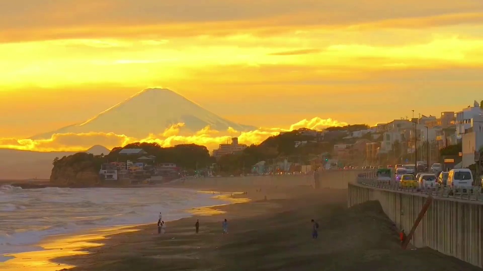 写实海滨风光画面，锥形积雪远山被暖黄阳光照射，暖金色夕照铺洒在沿岸沙滩、海浪、城镇建筑与沿海公路上。