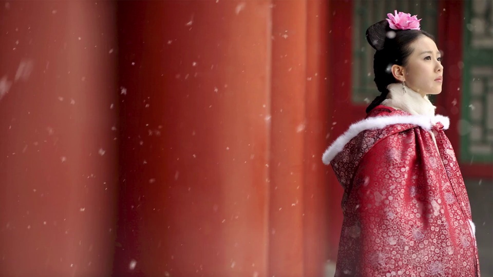古装女子立在飘雪的红色古建旁，身着带白边的暗纹红披风。