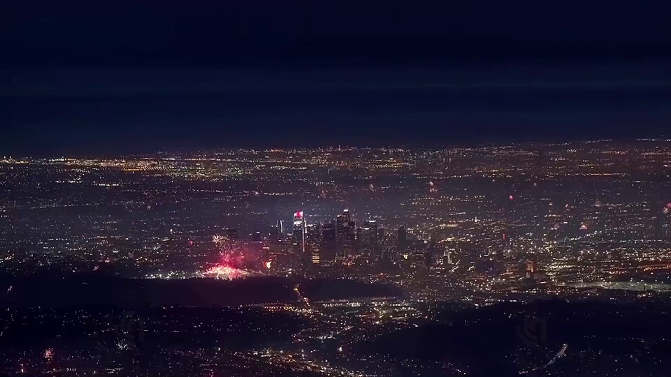 高空俯瞰的城市夜景，连片暖调灯火间点缀多处盛放的烟花