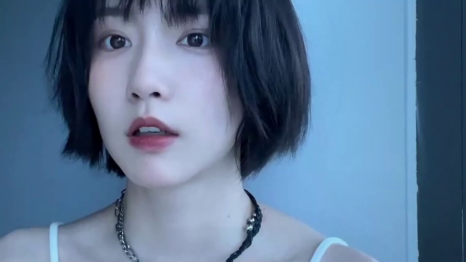 留黑色齐耳短发齐刘海的女性成果近景，穿白色细吊带，佩戴银链配黑编绳颈链，背景为冷调浅蓝色纯色墙面。