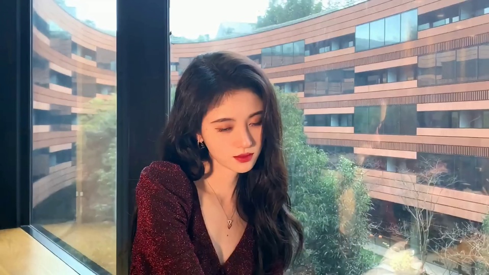 靠窗位置，身着带银白细闪的酒红色V领服饰的年轻女性低垂眼睑，背景窗外是带棕红横向线条的现代建筑与绿树，窗边露出浅木色桌面