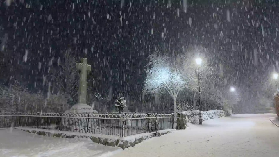 雪夜户外步道，黑色铁艺围栏后方立着石质十字纪念碑，旁侧是挂满积雪的无叶小树，沿线路灯散发出暖白色光线，地面、围栏与石墙都积着厚雪，背景是深暗的落雪夜空。