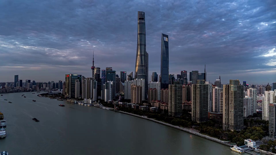 黄浦江江面与对岸陆家嘴摩天楼群组成的城市天际线，沿江岸分布着深灰绿色树木，画面整体呈灰蓝色冷调。