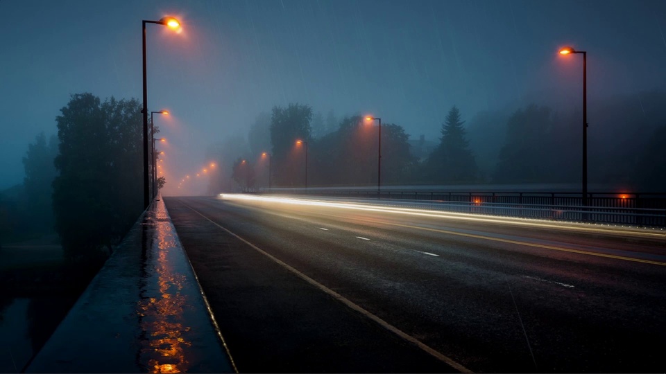 雨雾笼罩的雨夜公路桥，暖橙色路灯沿路延伸，湿滑路面映着光线与长曝光拖光