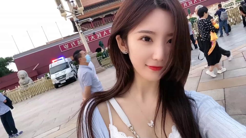 长发年轻女子在红墙中式建筑前自拍，广场上有行人、白色警车和石狮子。