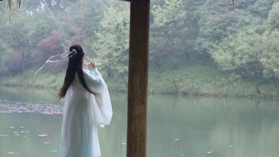 雨雾中湖亭下，身着浅蓝白渐变广袖古风汉服的女子背对镜头横吹白色长笛，前景湖面浮着荷叶碎影，背景是雾气笼罩的葱郁山林。