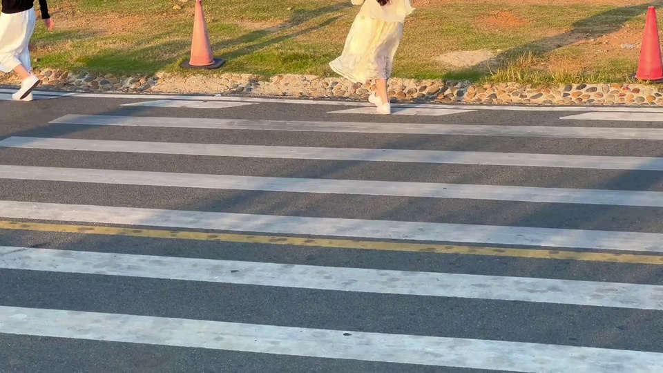 黄昏户外斑马路上，白裙女孩背对镜头向前行走，路侧草地上立有橙色交通锥