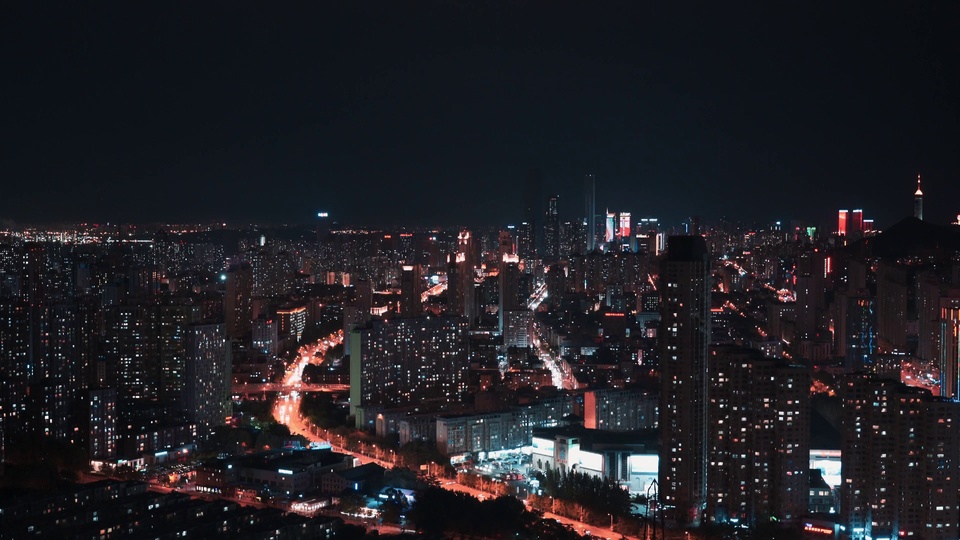 高空俯视视角下的灯火城市夜景，连片楼宇错落分布，道路上的车流延伸出暖橙色光轨。