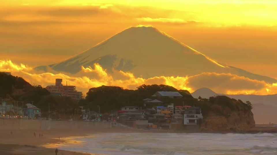 暖橙日落天光下，海浪轻拍沙滩，远处锥形雪峰衬着沿岸的滨海小镇与金霞。