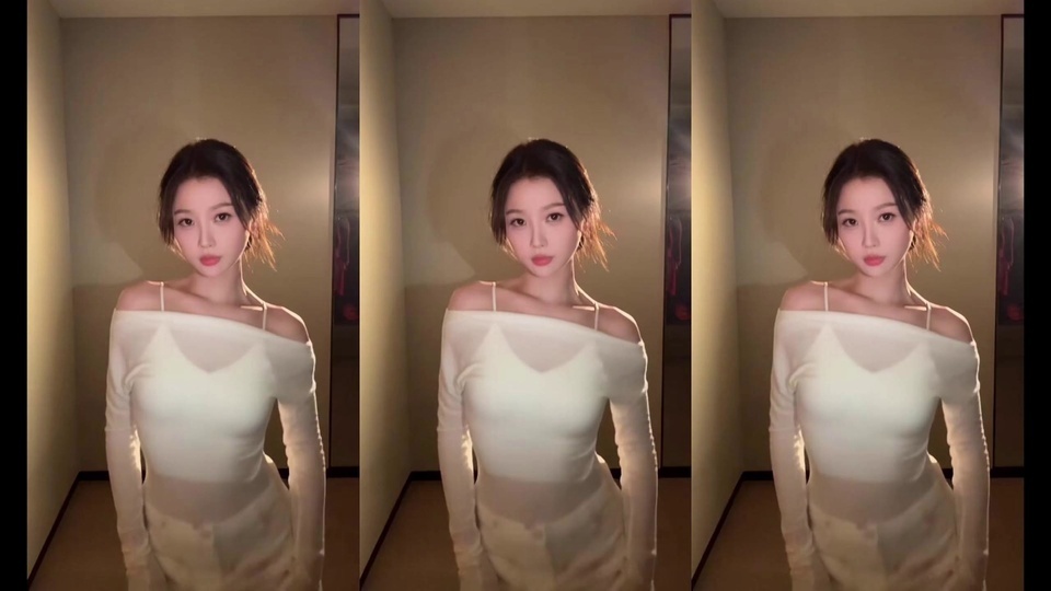 深色盘发的年轻女性身着米白色露肩细肩带长袖上衣，站在暖光笼罩的室内走廊中，转头望向侧方，造型温婉柔和。