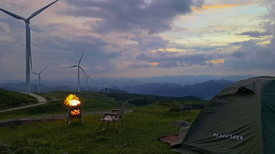 黄昏山地草甸露营场景，青绿色草地上摆放着露营帐篷、燃着橙黄色火焰的炉具与折叠露营椅，背景是淡紫灰色黄昏天空。
