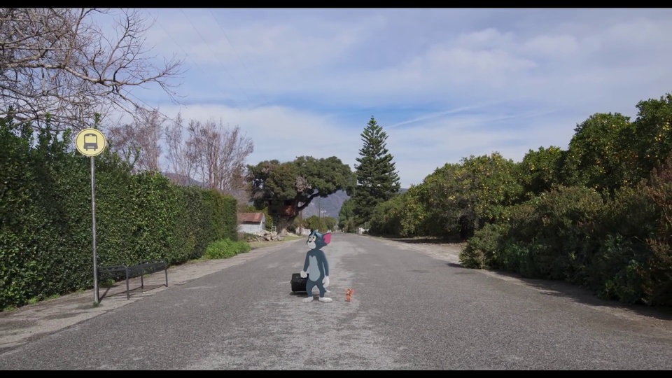 郊外郊外沥青公路边，灰蓝色拟人卡通猫站在黑色行李箱旁，脚边立着橙色小老鼠卡通形象，周边分布绿篱、公交站牌与金属长椅。