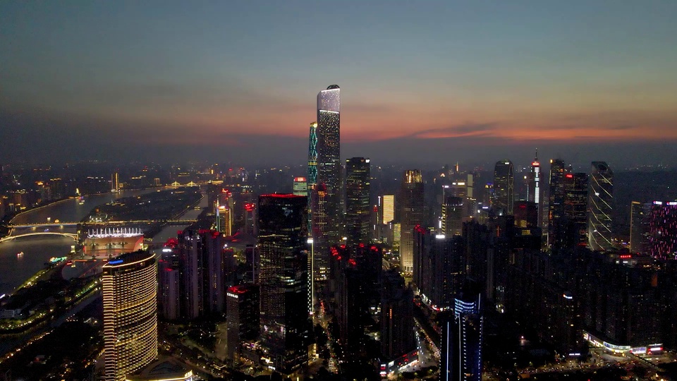 暮色黄昏中的城市江景航拍，高楼林立灯火璀璨，远处天际晕染橙红霞光。