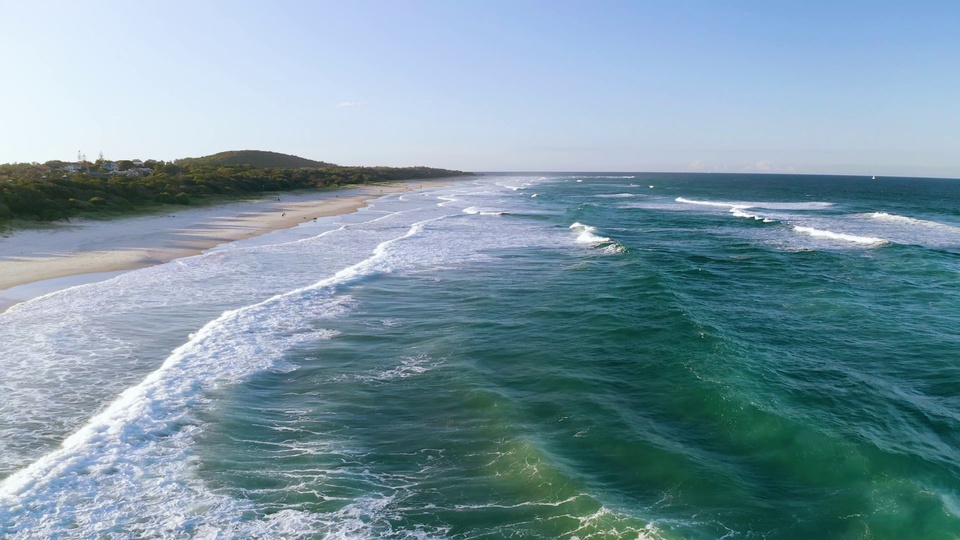航拍视角的晴朗海滨海景，米黄色沙滩向远方延伸，青绿色海浪带着白色浪沫层层涌向岸边，上方是明净的浅蓝晴空。