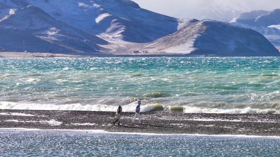雪山下的湖畔碎石滩上，两名行人踏滩前行，浪涛拍岸