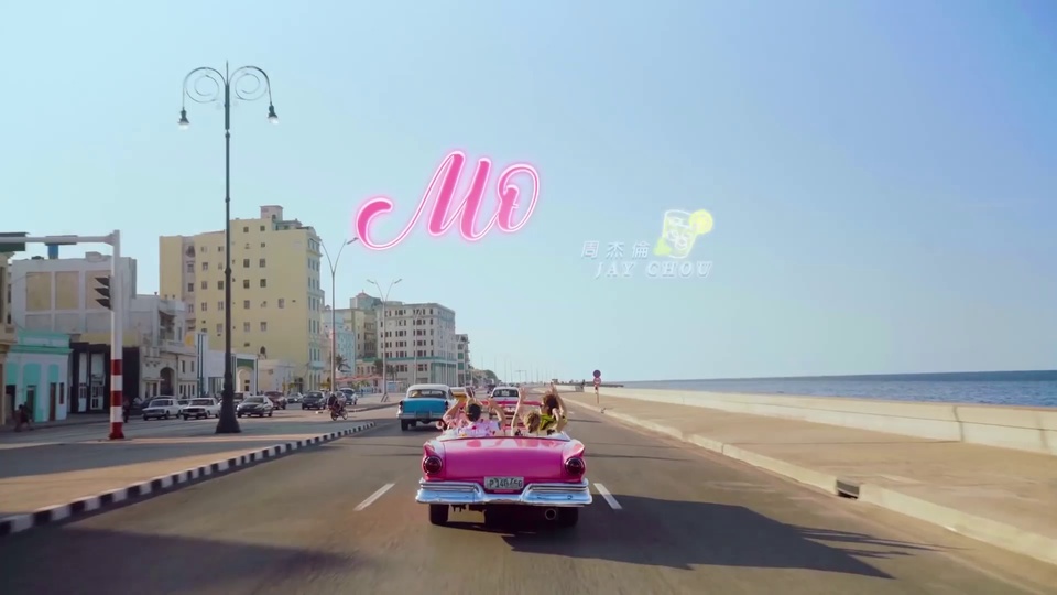 晴朗日间的滨海城市道路场景，亮粉色敞篷车向前行驶，旁侧车道有蓝色复古轿车，道路左侧是浅色复古临街建筑，一侧衔接开阔海面与浅蓝晴空。