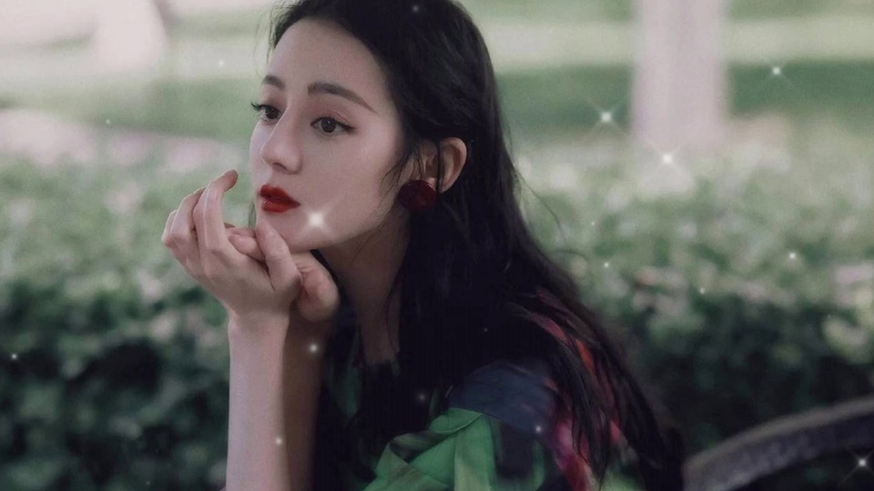 年轻女性留黑色长卷发，戴红色圆形耳饰、涂正红色口红，穿绿底带红黑花纹上衣，双手托腮望向画面左侧，背景是虚化的绿色灌木丛，散布细碎白色星芒光斑。