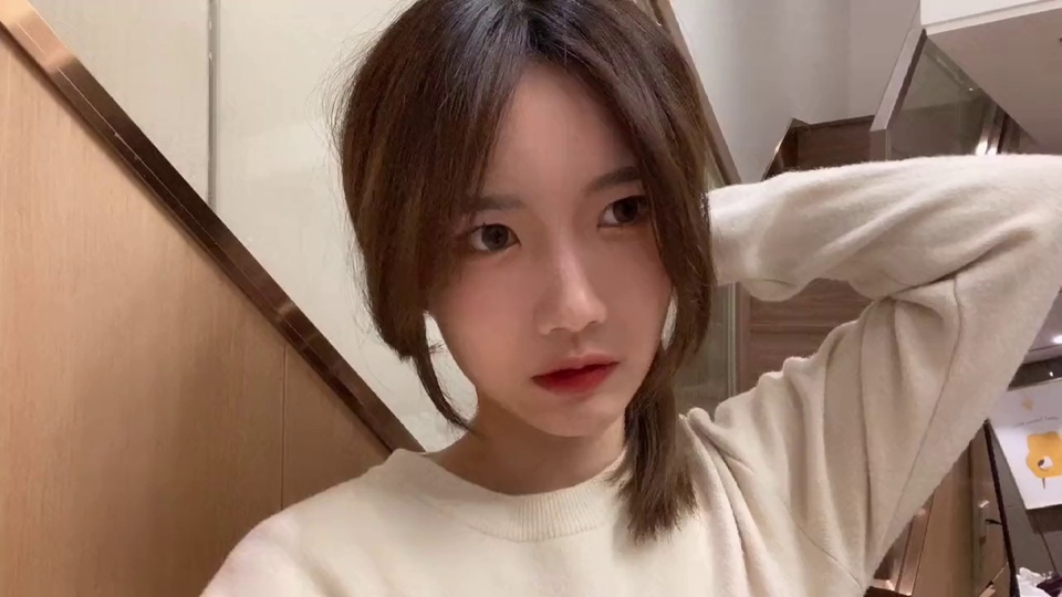 室内楼梯旁，作为美女、黑丝主题的年轻女性留深棕色齐肩碎发，身着米白色圆领长袖上衣，左臂抬至头后，背景为浅木色楼梯侧板与白墙，暖调柔和。