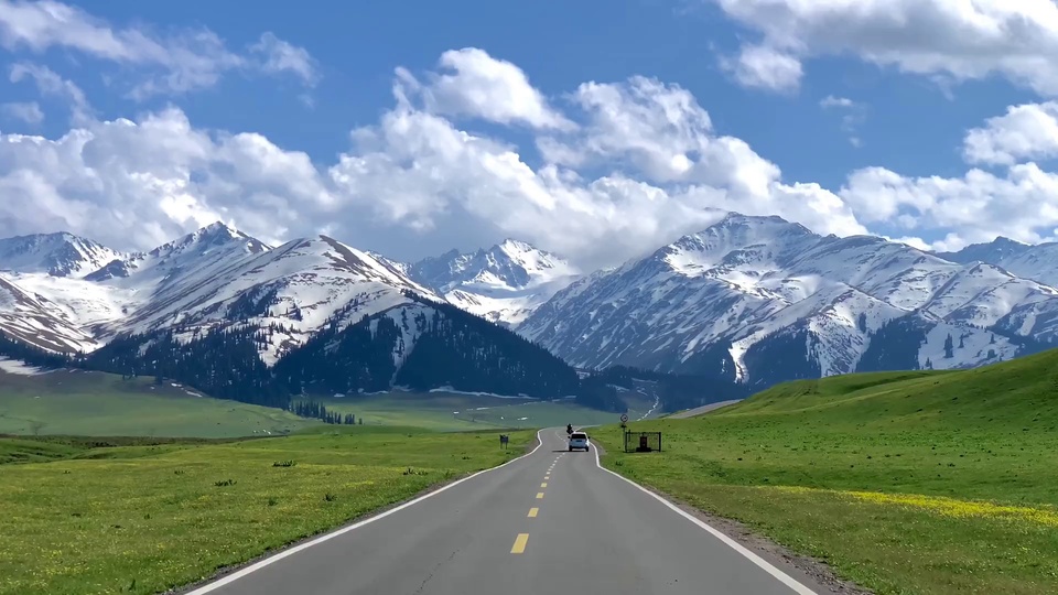 写实摄影风格的草原雪山公路画面，深灰色柏油公路向远方延伸，两侧是翠绿草坡与针叶林，远处矗立着覆雪的连绵山脉，上方是飘着白云的澄澈蓝天。